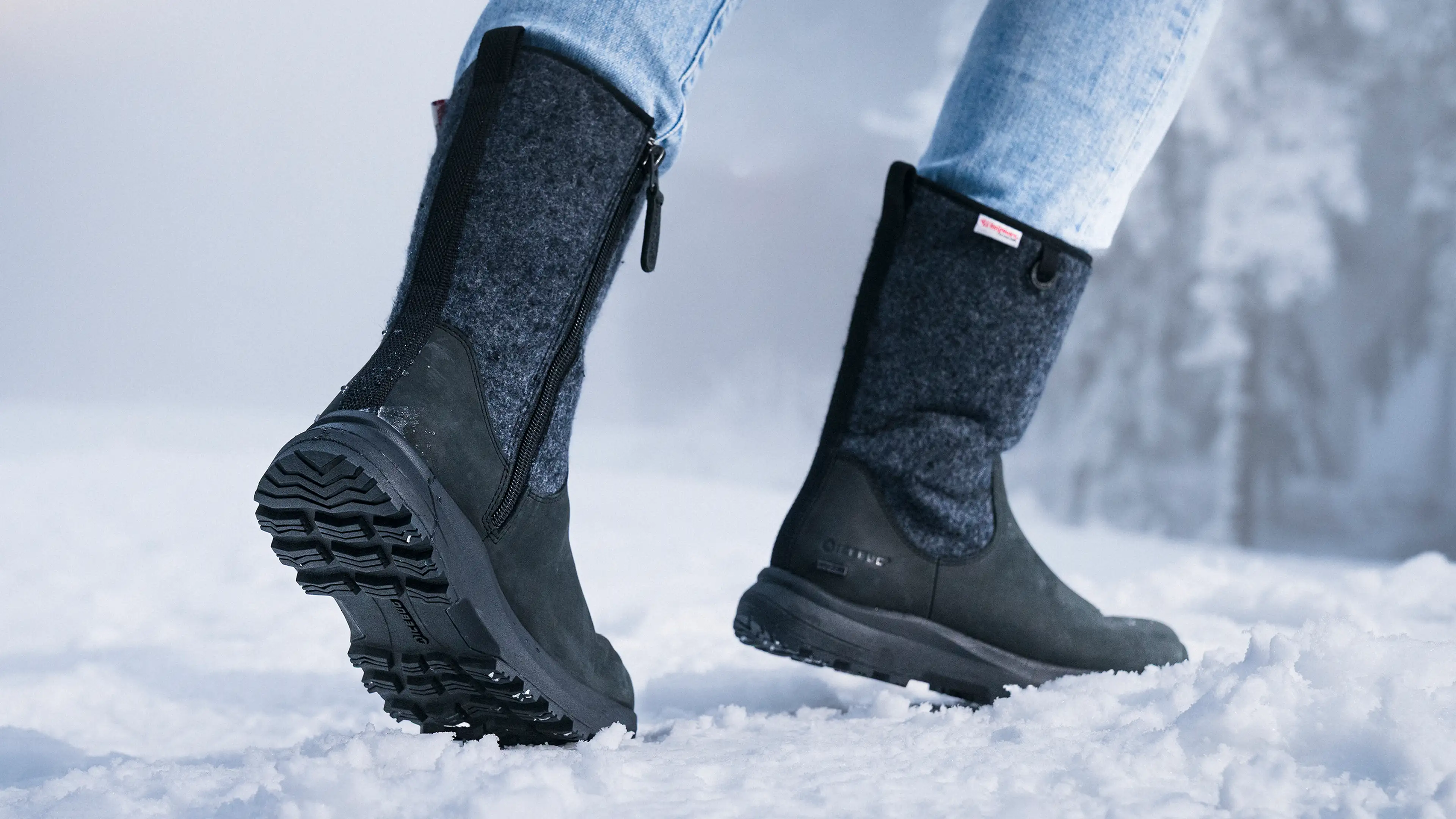 Close-up of a person wearing black winter boots and blue jeans, walking on snow. The boots have textured soles and a side zipper.