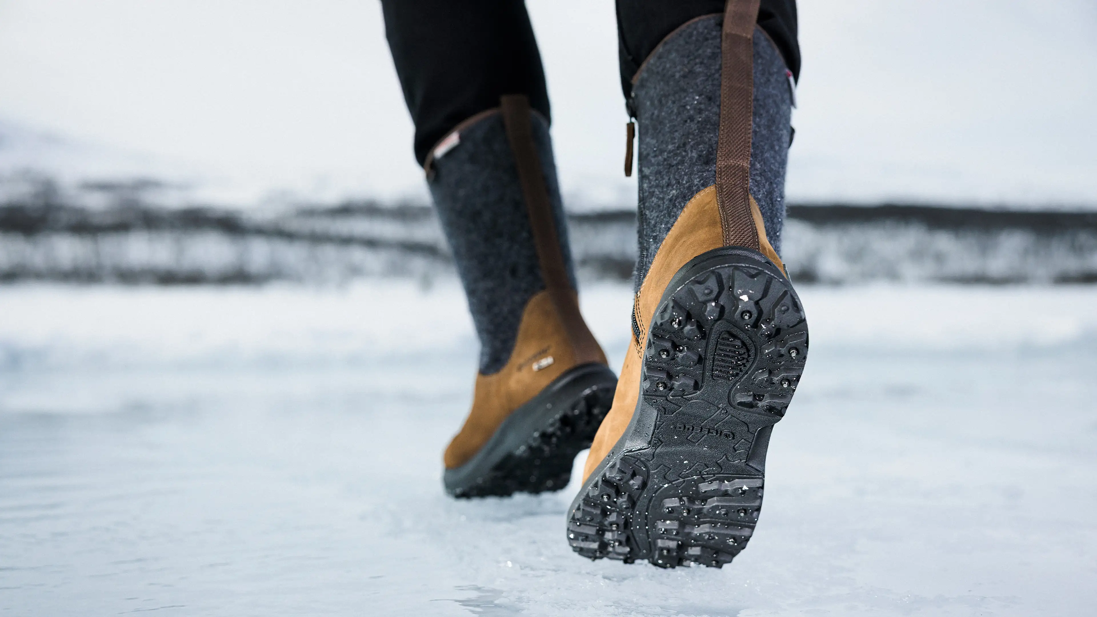 Close-up of a person walking on ice, wearing brown winter boots with studded soles, set in a snowy landscape.