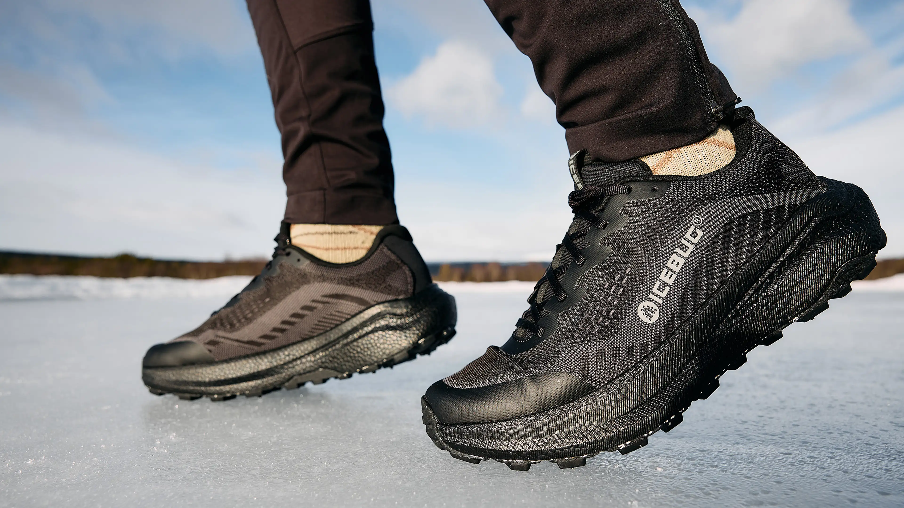 Close-up of a person wearing black Icebug running shoes on a frozen surface, with mountains and a cloudy sky in the background.