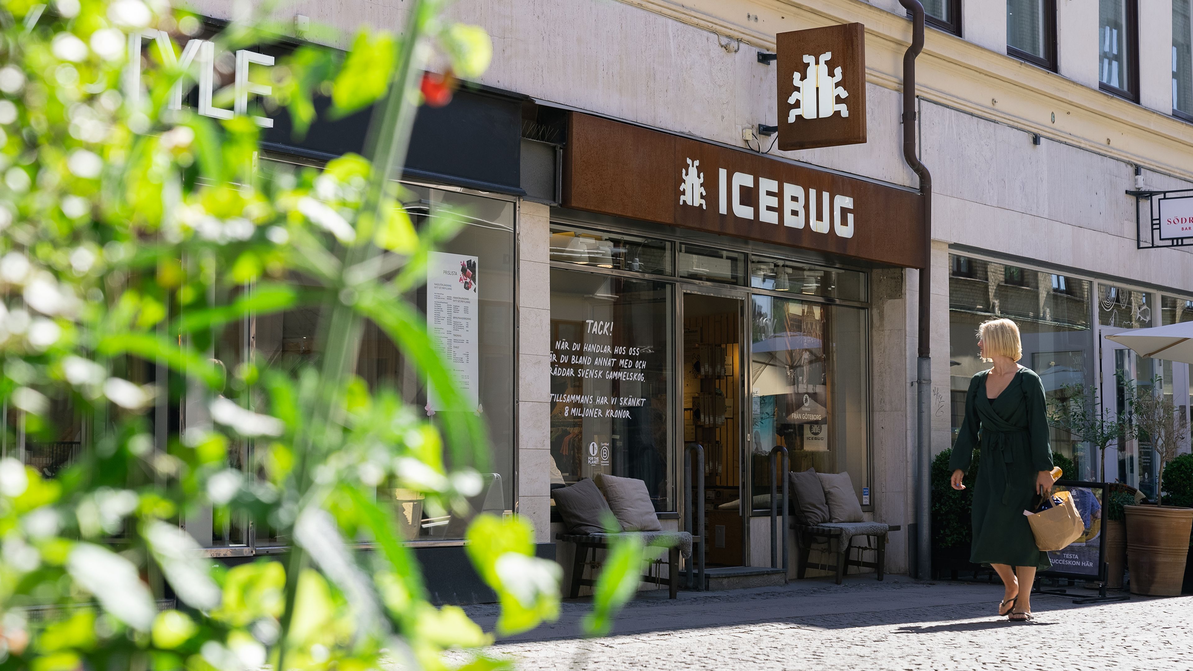 A woman walks past the Icebug store on a sunny day, with green plants in the foreground and outdoor seating visible.
