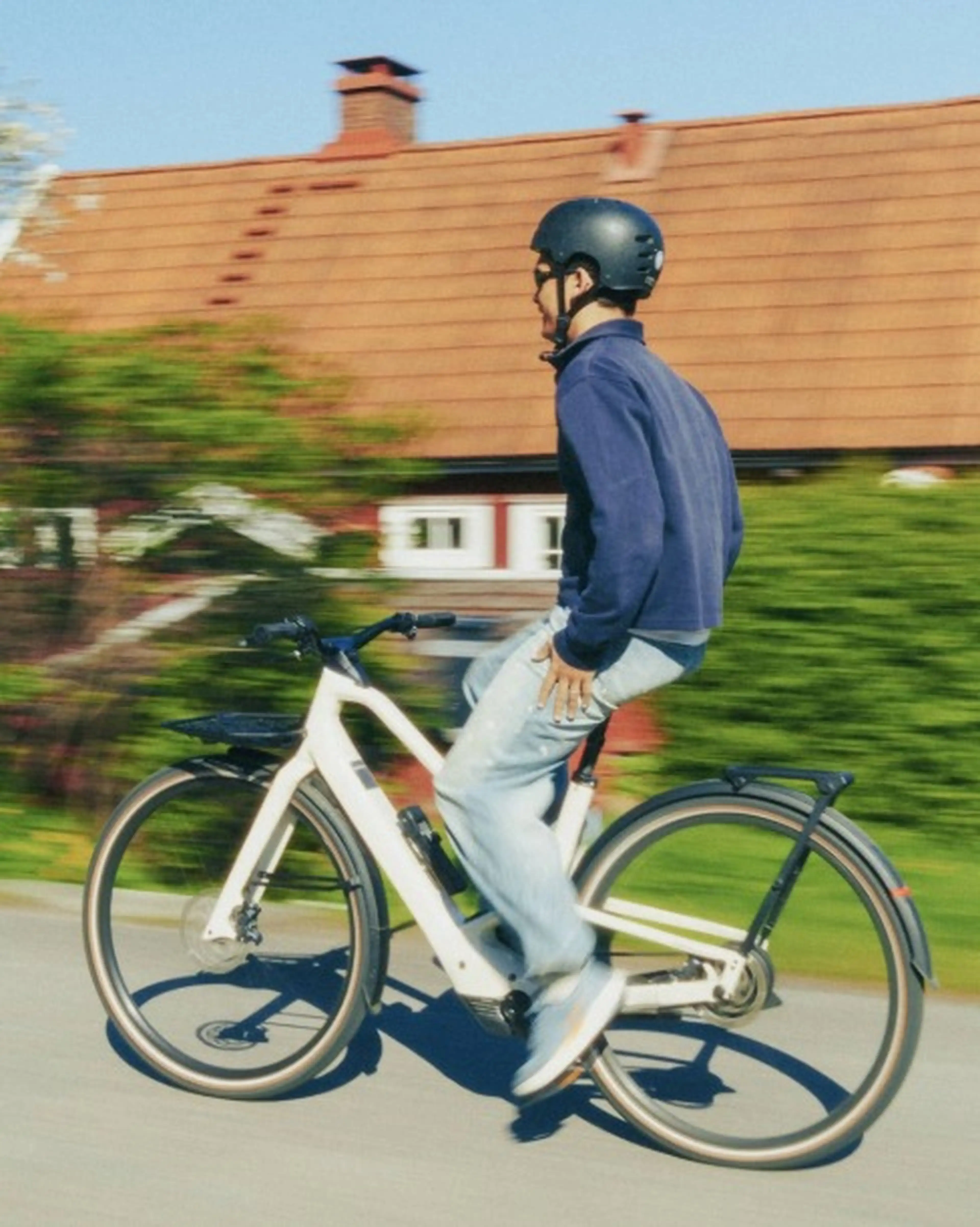 Person wearing a helmet rides a white bicycle past a house with a red roof on a sunny day.