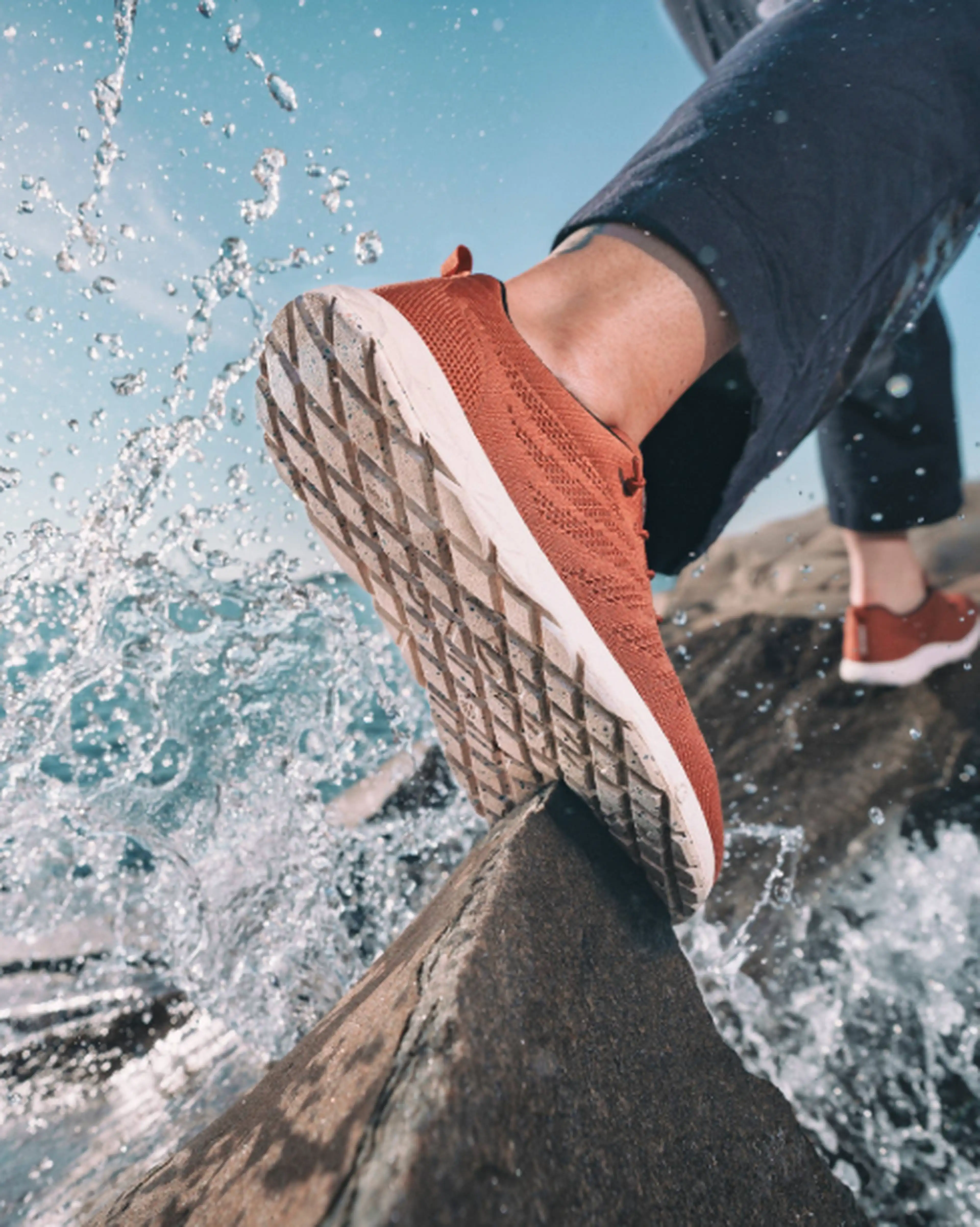 Close-up of a person wearing red sneakers stepping on a wet rock with water splashing, against a blue sky background.