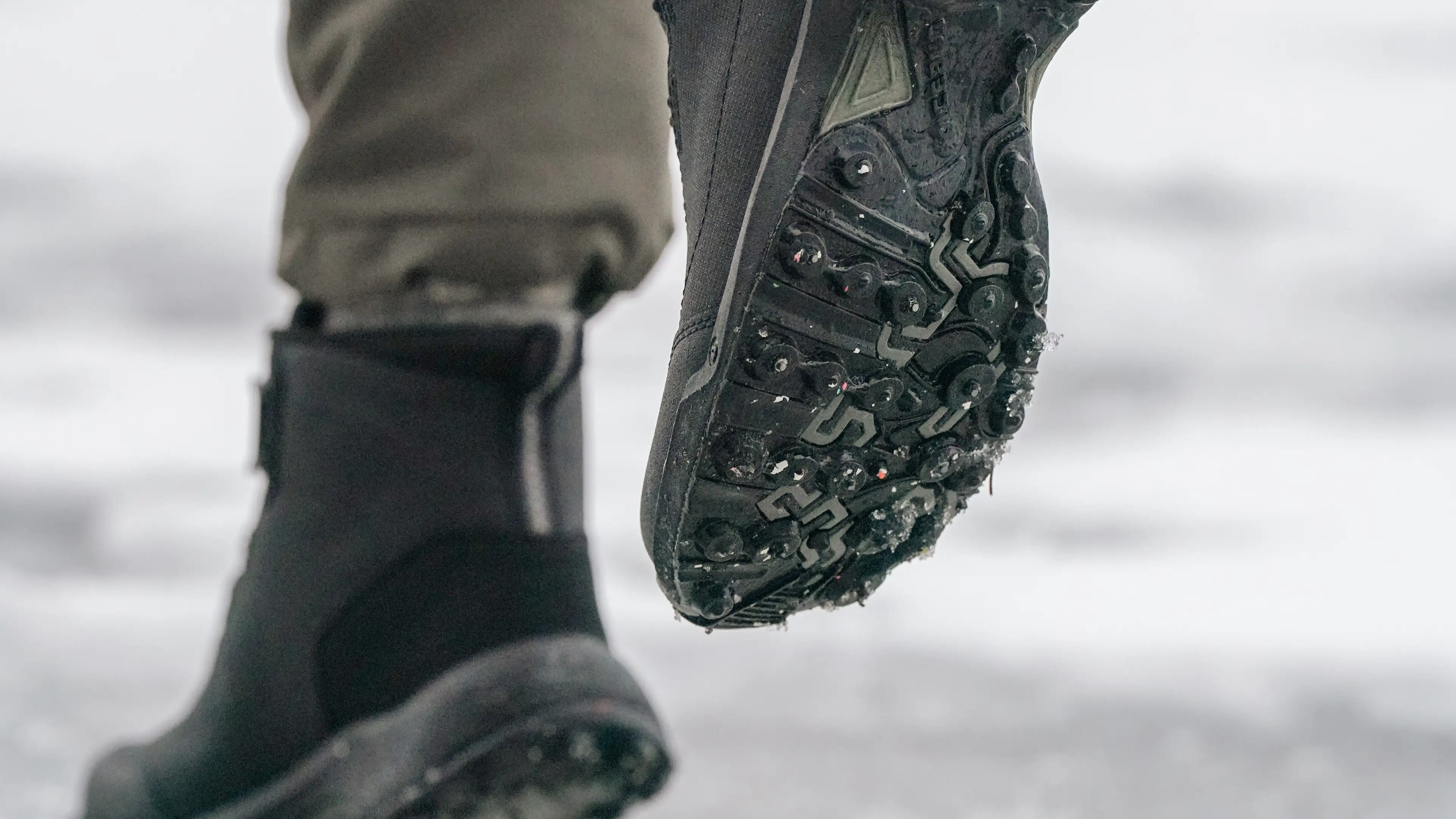 Close-up of a person walking on ice, wearing studded Icebug boots with cleats for traction. The ground appears snowy and slippery.