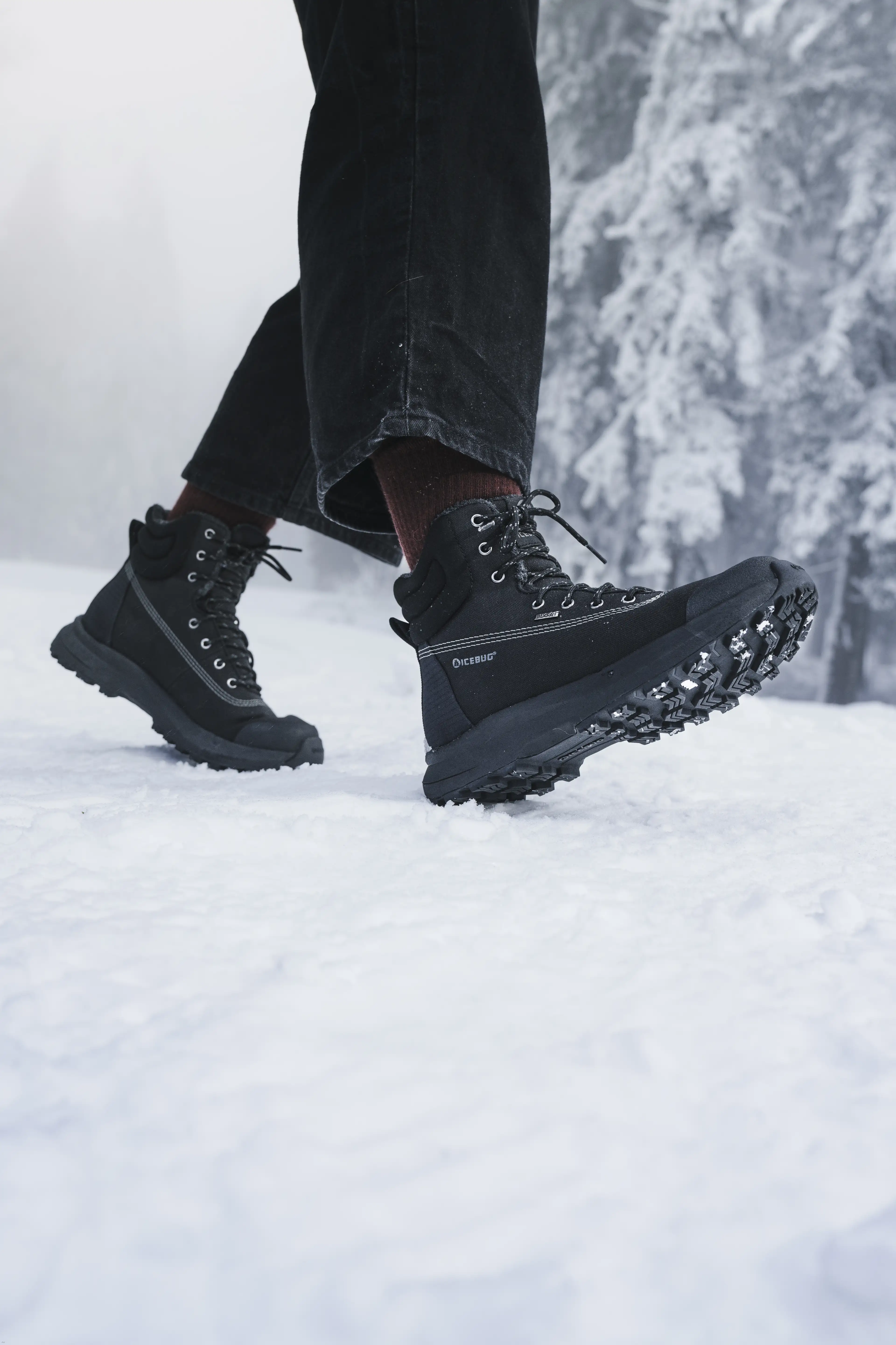 Person wearing black hiking boots walking on snow with snow-covered trees in the background.