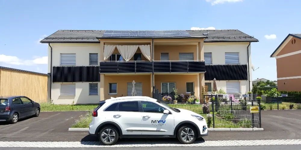 Four-unit house with rooftop PV panels, garden, and parking spaces; a my-PV car is in the foreground.