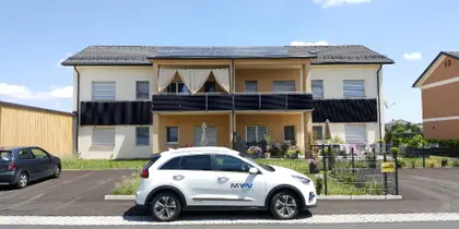 Four-unit house with rooftop PV panels, garden, and parking spaces; a my-PV car is in the foreground.