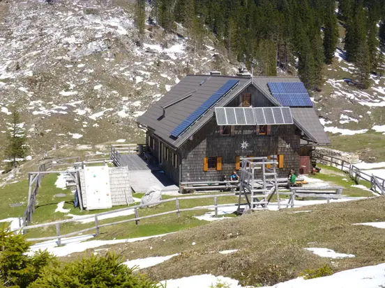 Rustic mountain cabin with solar panels on roof, surrounded by snow patches and evergreen trees.