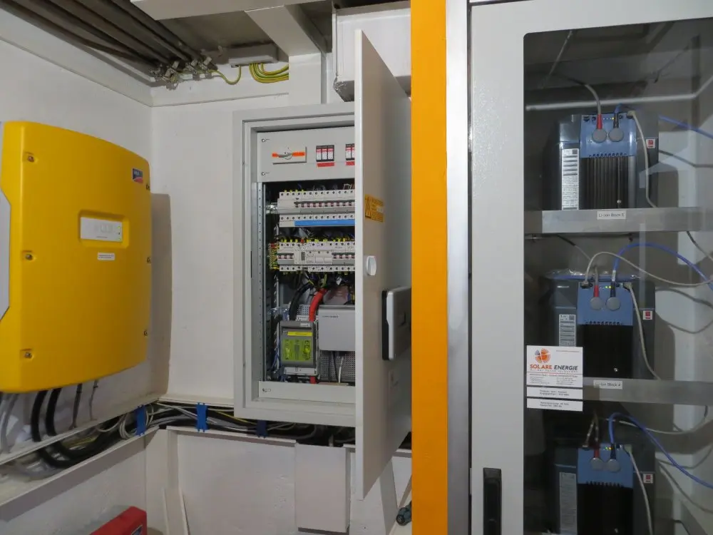 Electrical control room with open panel showing circuits, wires, switches; nearby batteries and yellow inverter visible.