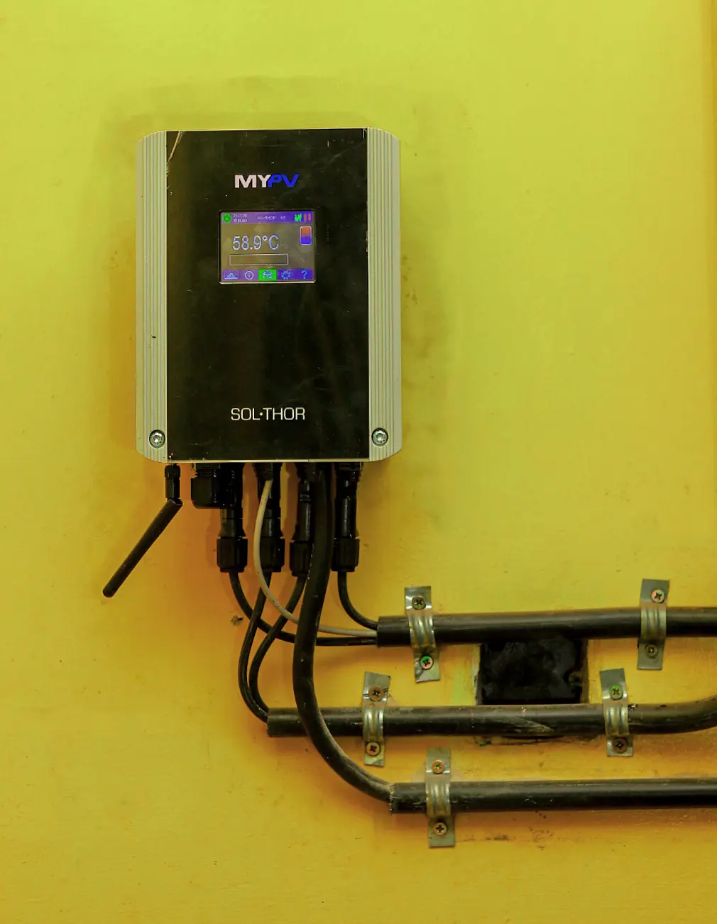 Wall-mounted digital thermostat with display showing "68.9°C," connected to four cables, set against a yellow background.