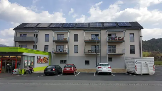 Modern three-story apartment building with solar panels, balconies, parked cars, next to a small green-yellow store