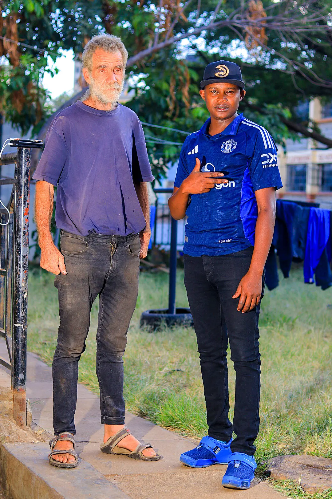 Two men standing outdoors; one in casual clothes and sandals, the other in a sports jersey and cap, both on a paved walkway with grass behind.