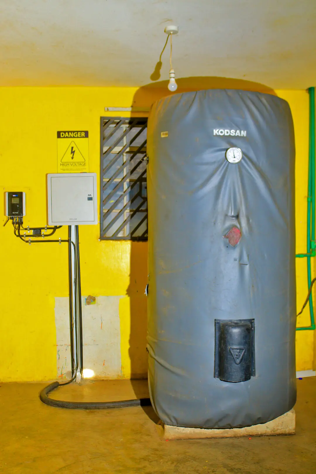 Large gray cylindrical water heater in a yellow room, labeled "KODSAN," with a nearby electrical panel and "Danger" sign.