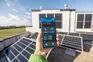 Hand holding a smartphone displaying a solar panel setup app, with solar panels on a rooftop in the background.