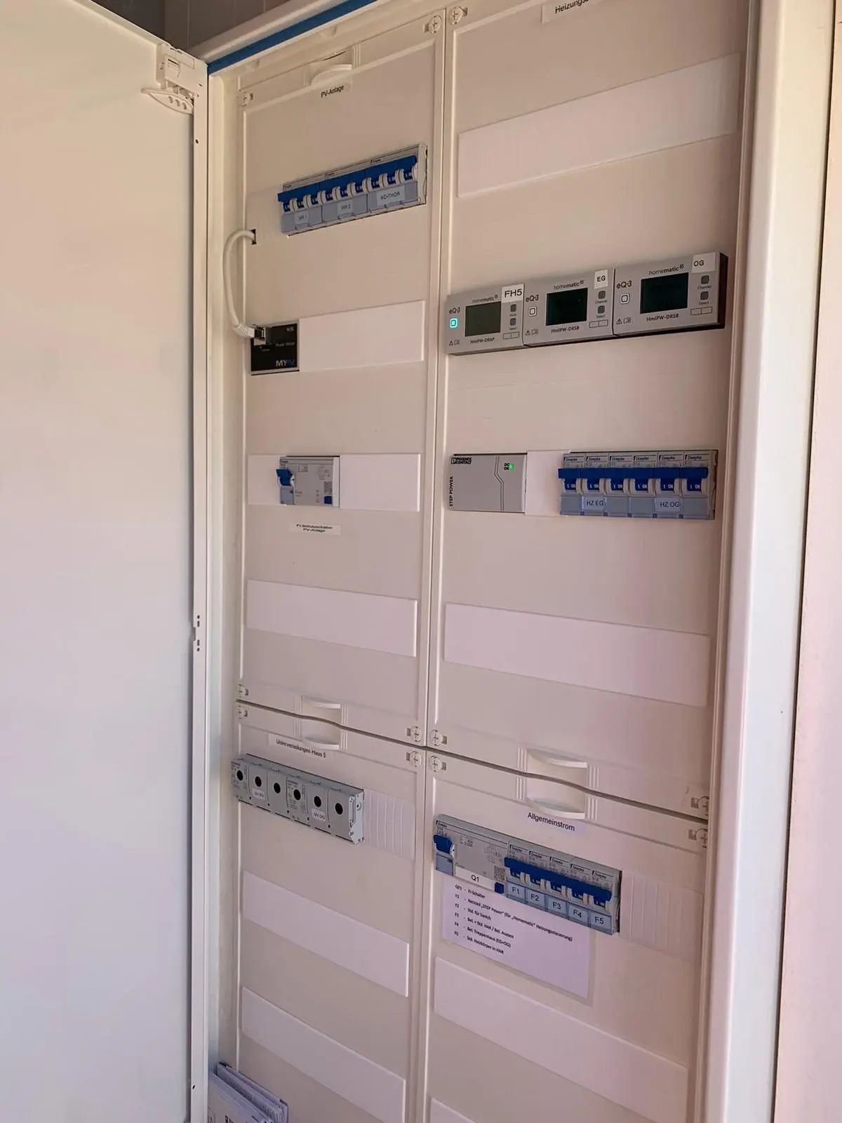 An open electrical panel with multiple circuit breakers, switches, and meters, organized in rows inside a white cabinet.