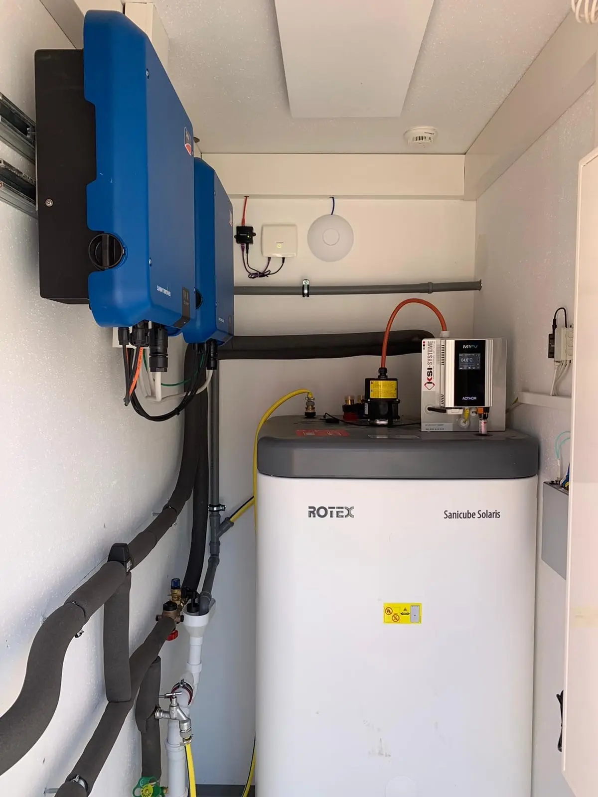 Small utility room with ROTEX Sanicube Solaris, AC•THOR, blue inverters, pipes, and electrical equipment on walls.