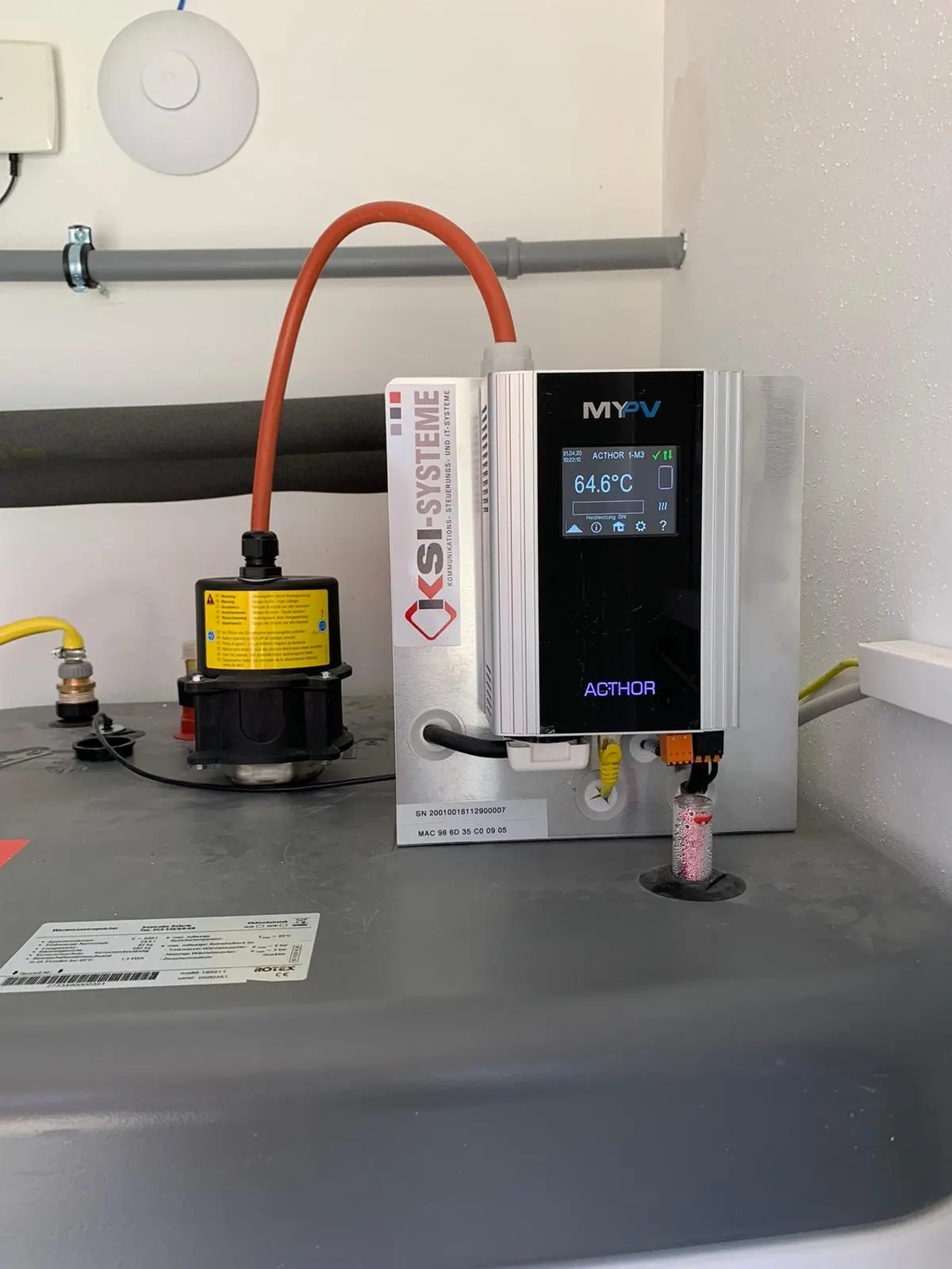 AC•THOR with digital display showing 64.6°C, connected to pipes and valves in a technical setup.