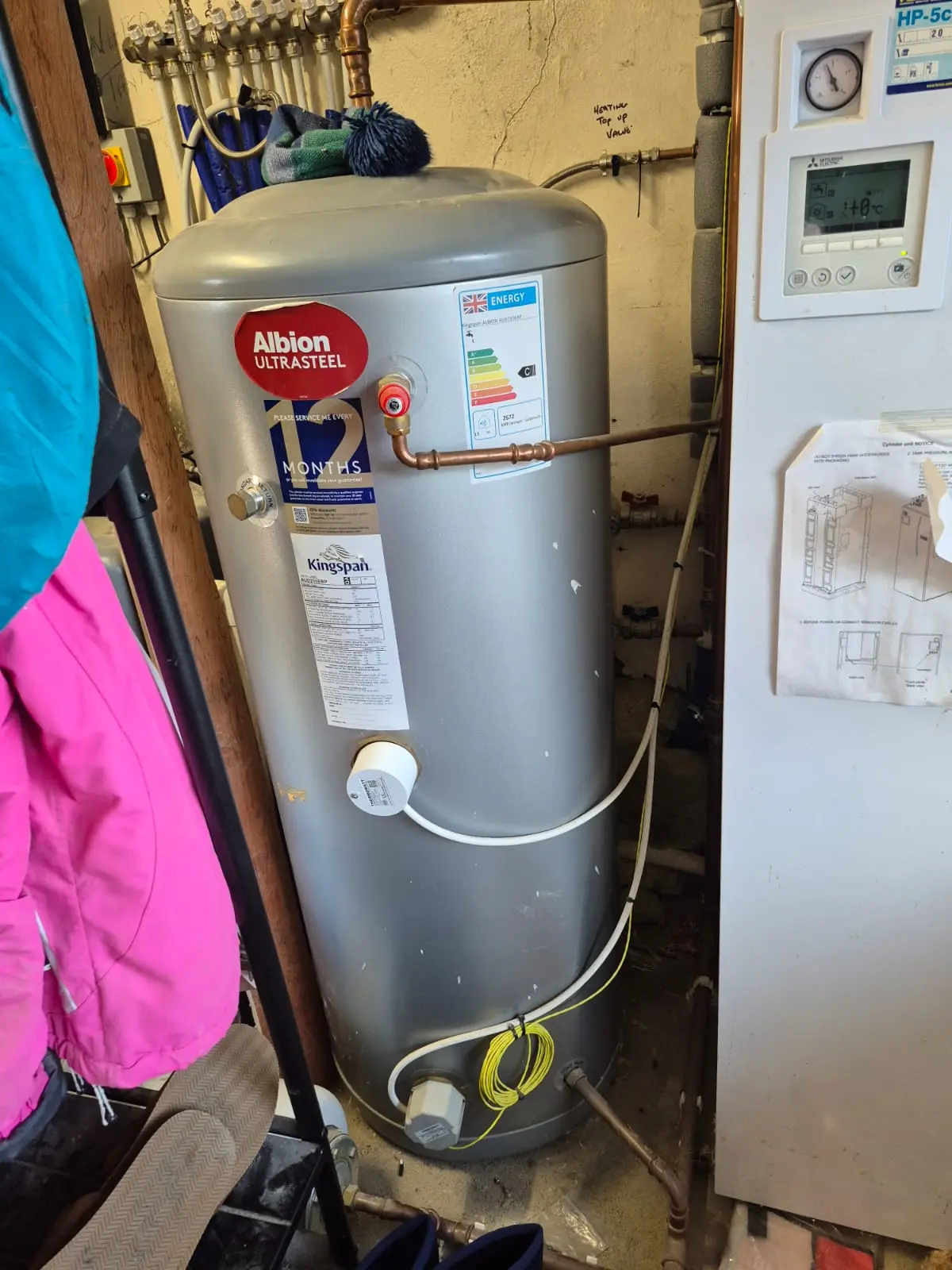 A silver water heater with labels and control panel is installed in a utility room, surrounded by colorful jackets and various items.