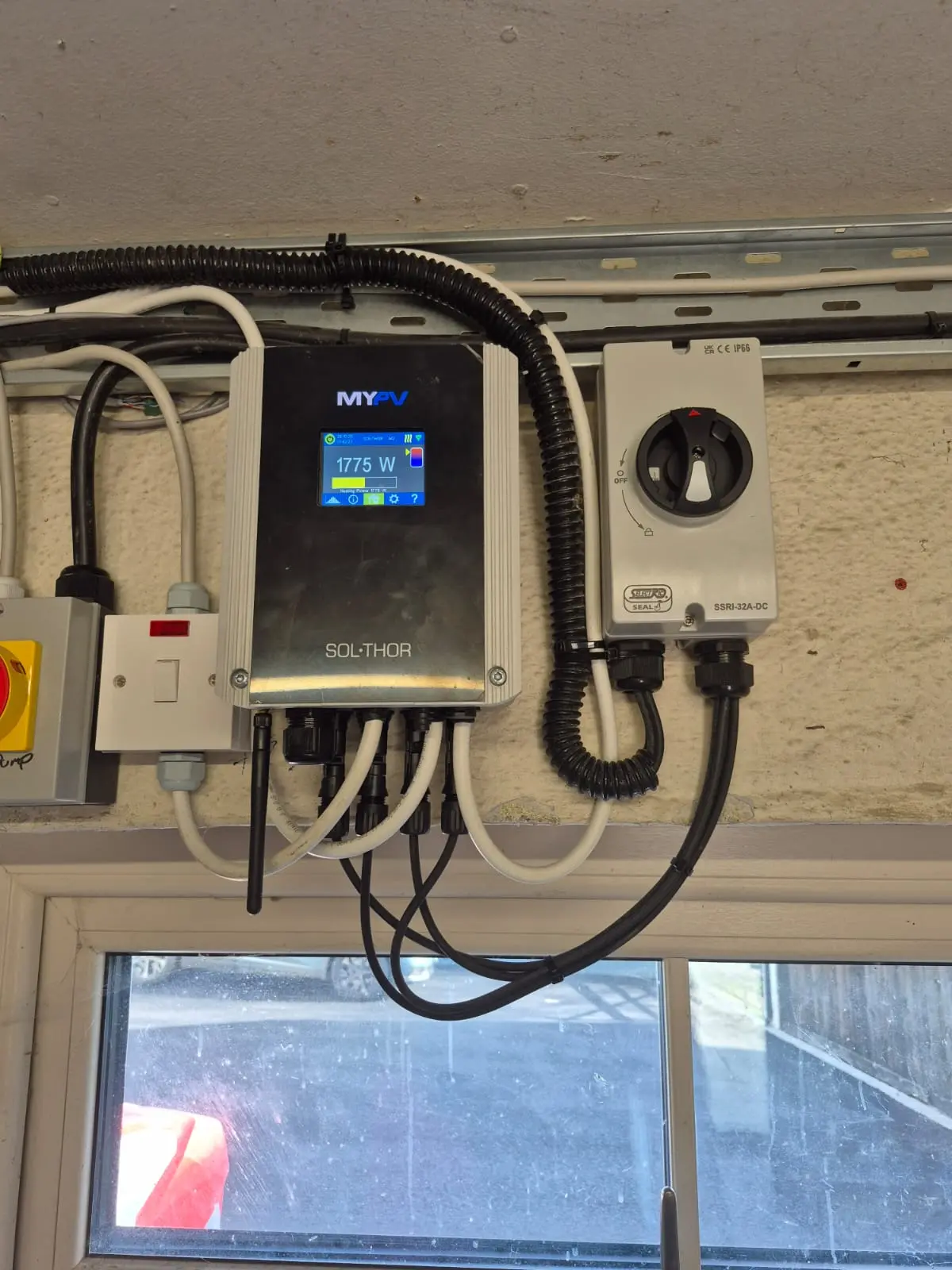 A power monitoring device with cables and wires is mounted on a wall above a window, displaying 1775 W on its screen.