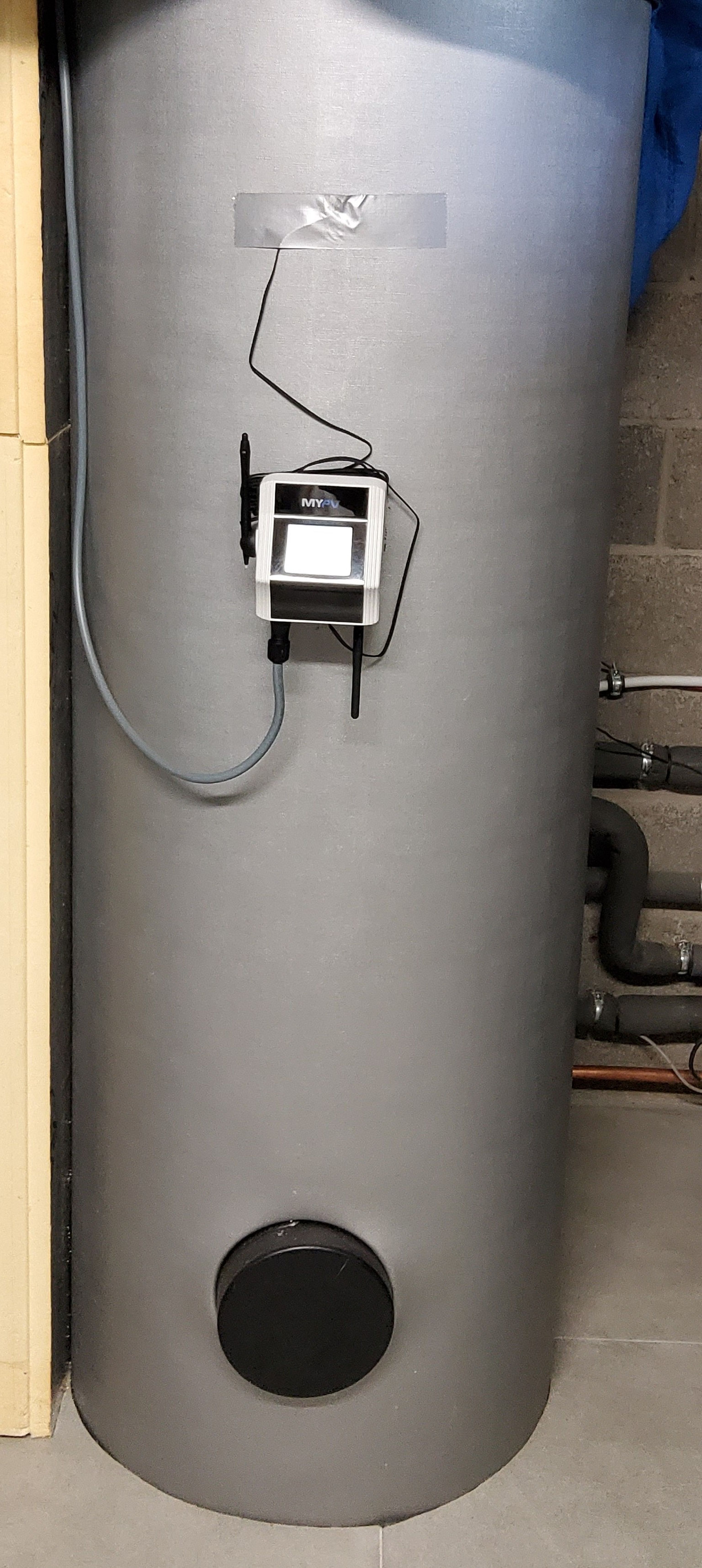 Large cylindrical metal water tank with AC ELWA 2 attached by wires, in a utility room with exposed pipes