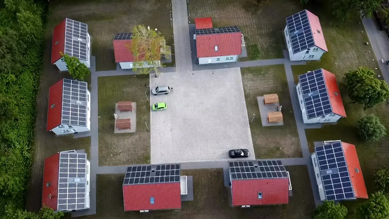 Aerial view of eight small red-roofed houses with solar panels surrounding a central paved area with parked cars.