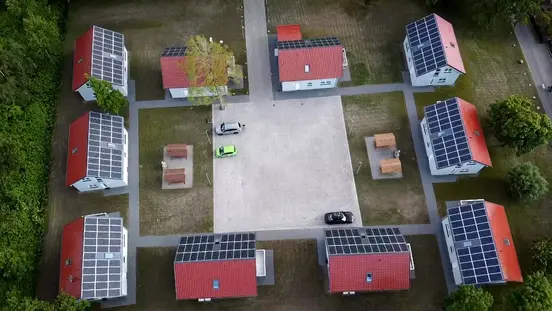 Aerial view of eight small red-roofed houses with solar panels surrounding a central paved area with parked cars.