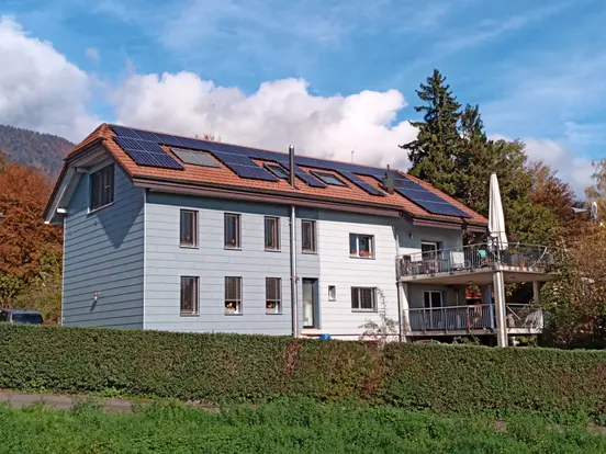 Two-story house with solar panels on the roof, surrounded by trees and greenery, under a blue sky with clouds.