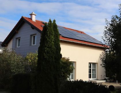 House with beige walls, red roof, and solar panels. Surrounded by greenery, clear sky above.