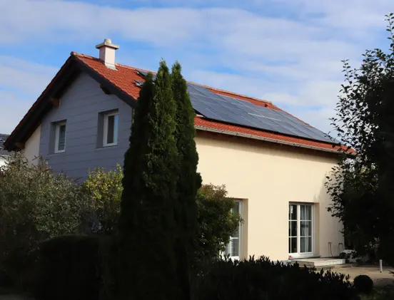 A house with a red roof, solar panels, and a beige exterior, surrounded by trees and shrubs, under a partly cloudy sky.
