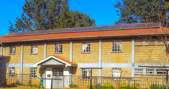 A two-story building with solar panels on the red roof, stone walls, multiple windows, and a person walking in front. Trees are in the background.