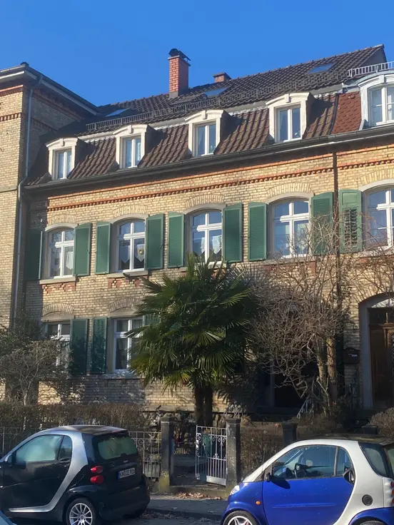 Renovated historic building with green shutters, palm tree in front yard, and two Smart cars parked in sunshine.