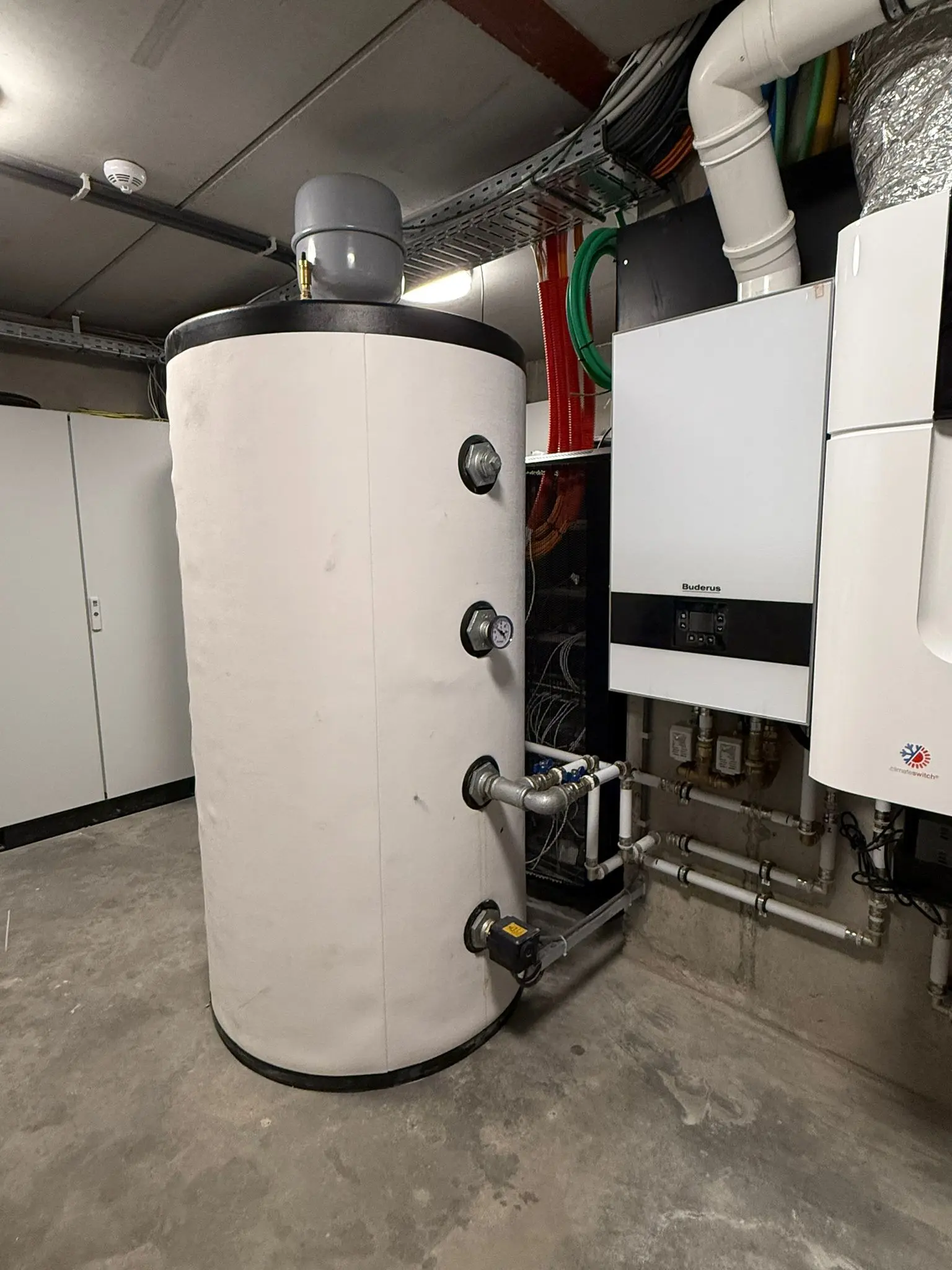 Large cylindrical water heater with gauges and pipes, connected to a wall-mounted boiler, in a utility room.