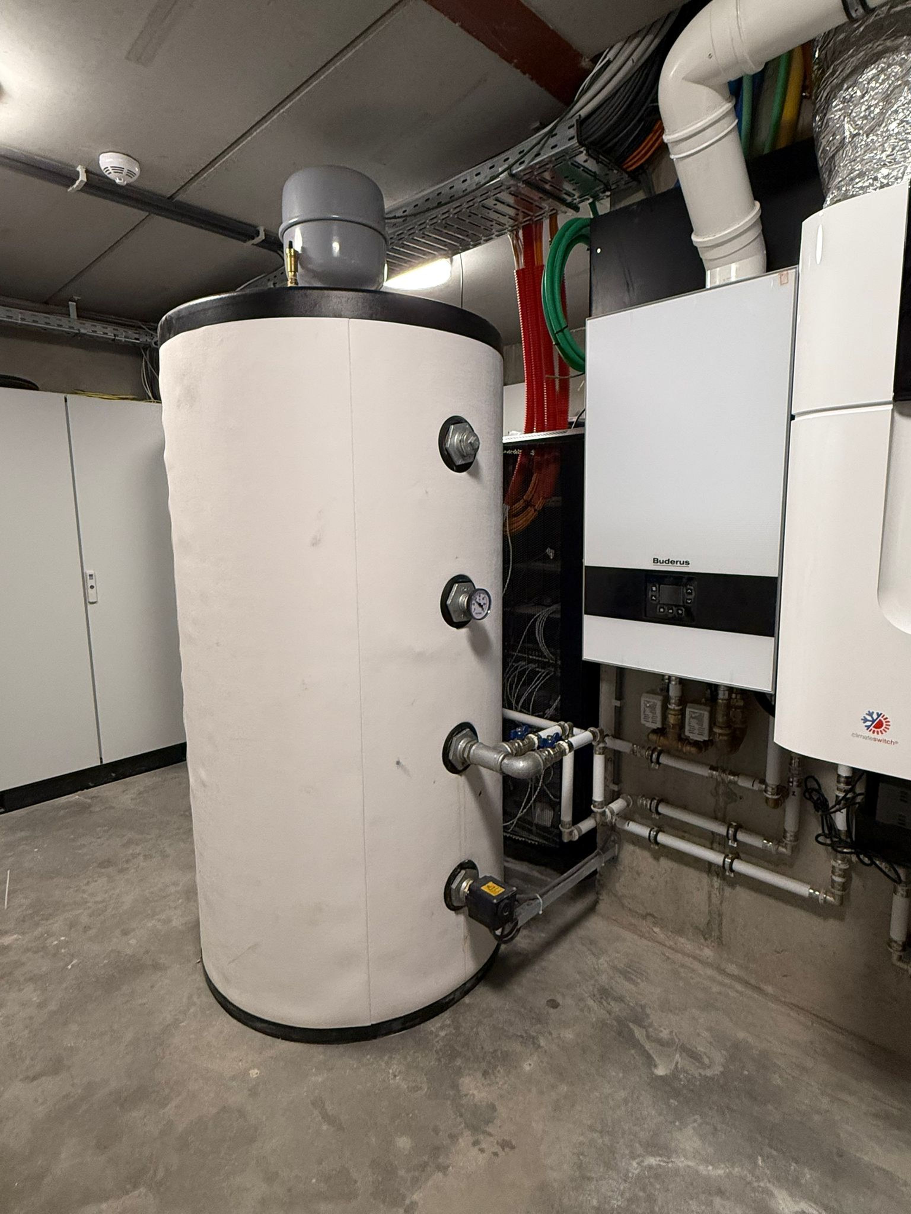 Large cylindrical water heater with gauges and pipes, connected to a wall-mounted boiler, in a utility room.