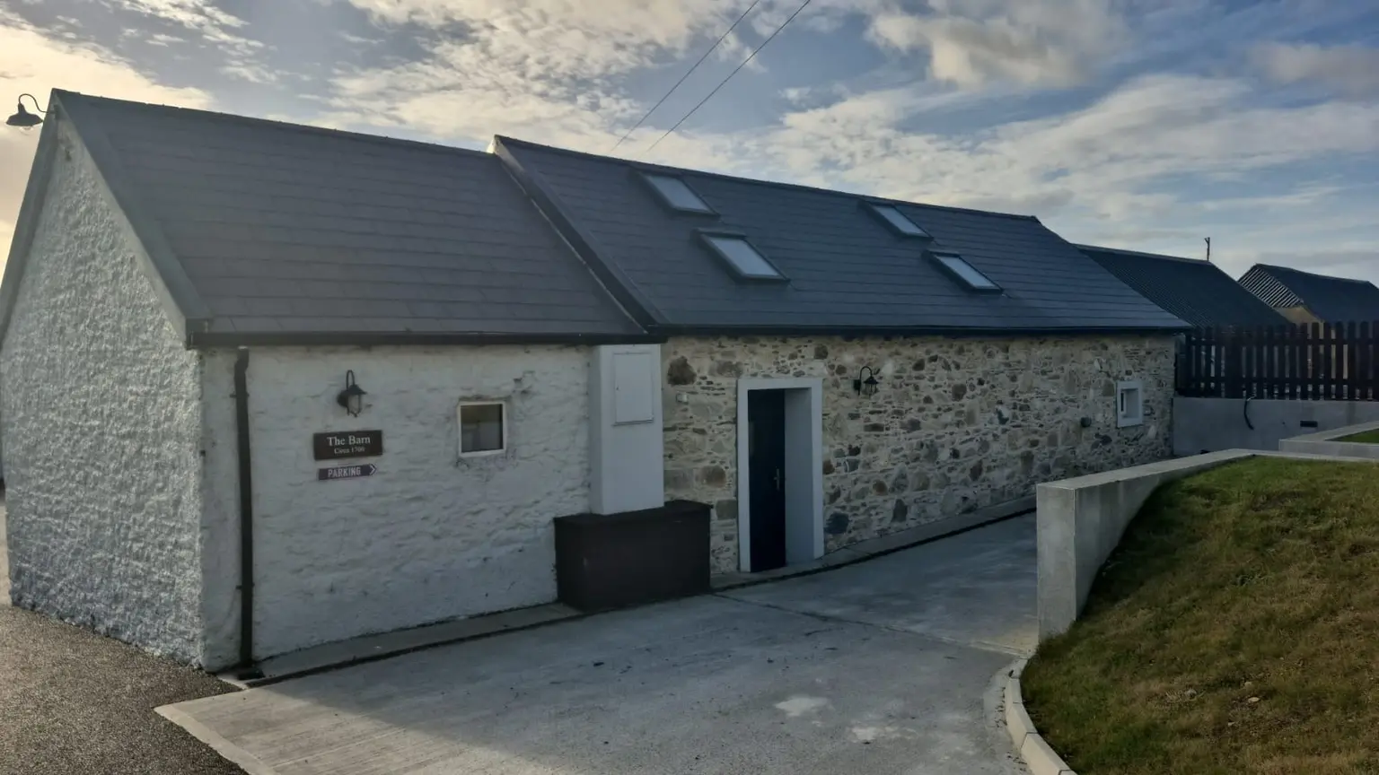 A stone cottage with a gray slate roof, several skylights, and a small sign by the door under a partly cloudy sky.