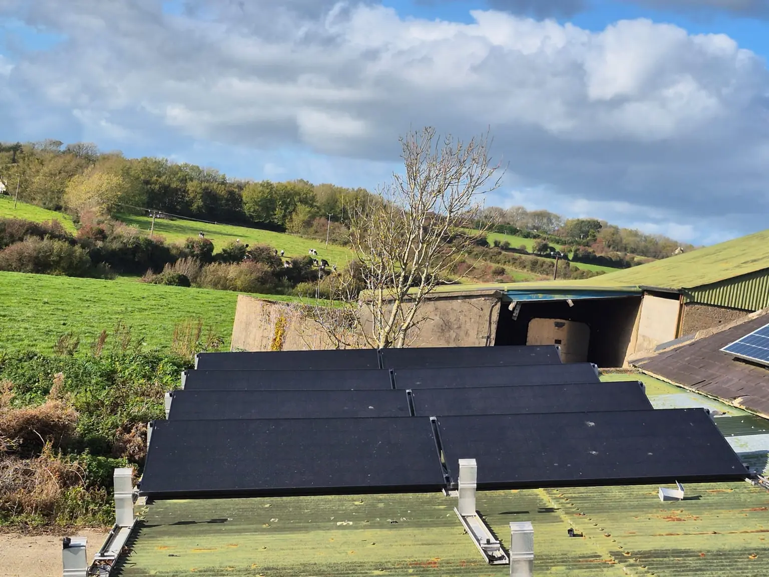 Solar panels on a rural building's roof surrounded by lush green fields and a cloudy sky background.