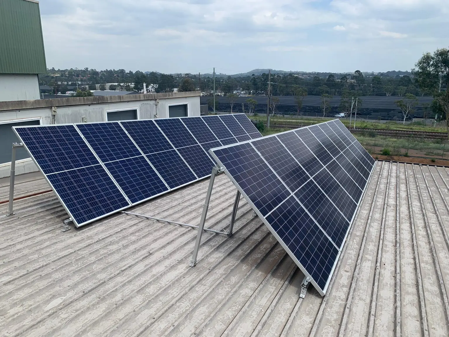 Solar panels on a corrugated rooftop under cloudy sky, with distant greenery and industrial buildings in view.