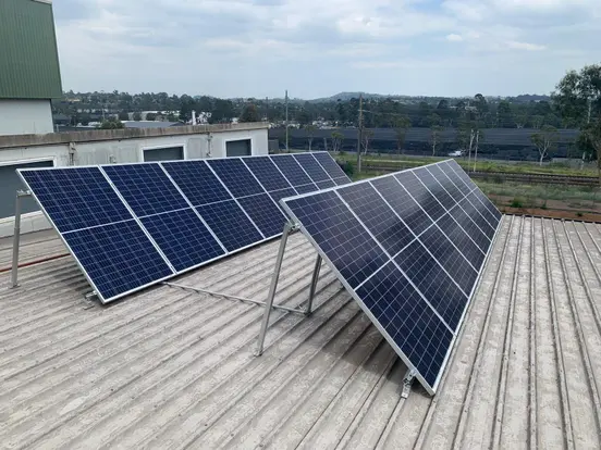 Solar panels on a corrugated rooftop under cloudy sky, with distant greenery and industrial buildings in view.