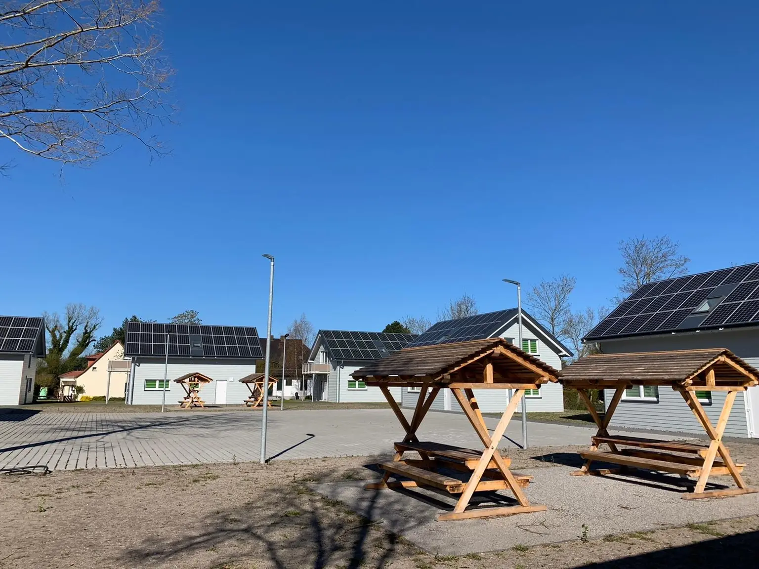 Row of modern houses with solar panels under clear sky, wooden picnic tables on paved area in foreground.