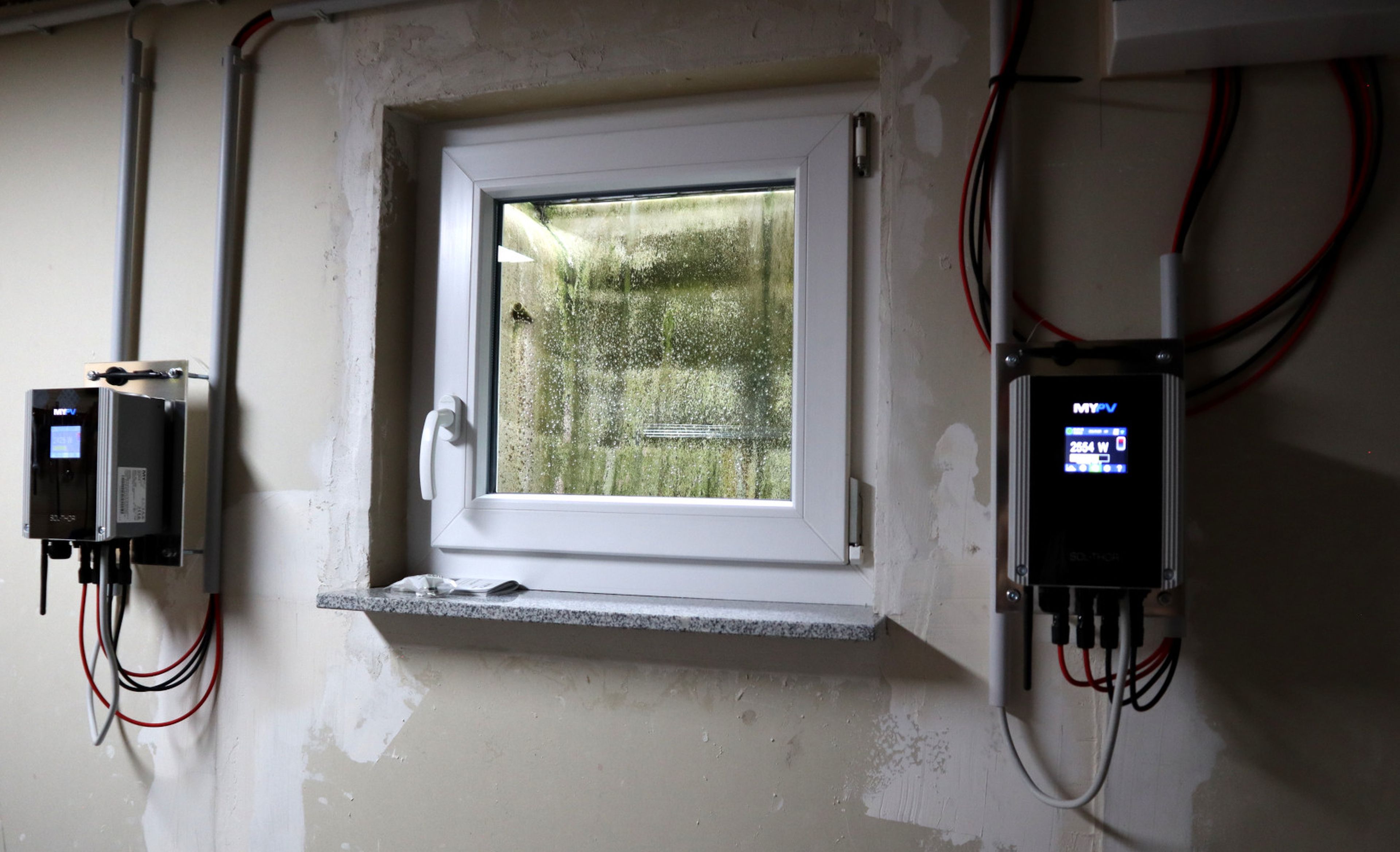 A small, square window in a basement wall with two electronic devices installed on either side, connected by wires.