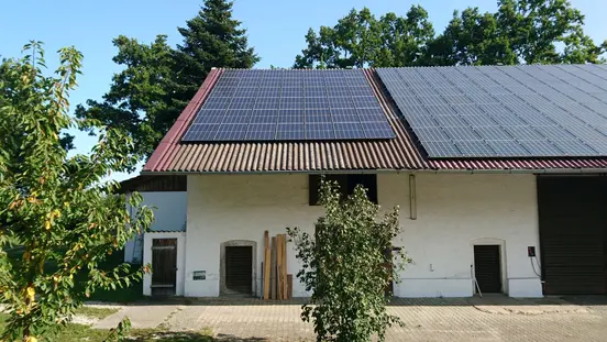 White building with sloped roof covered in solar panels, surrounded by trees and paved area in front.