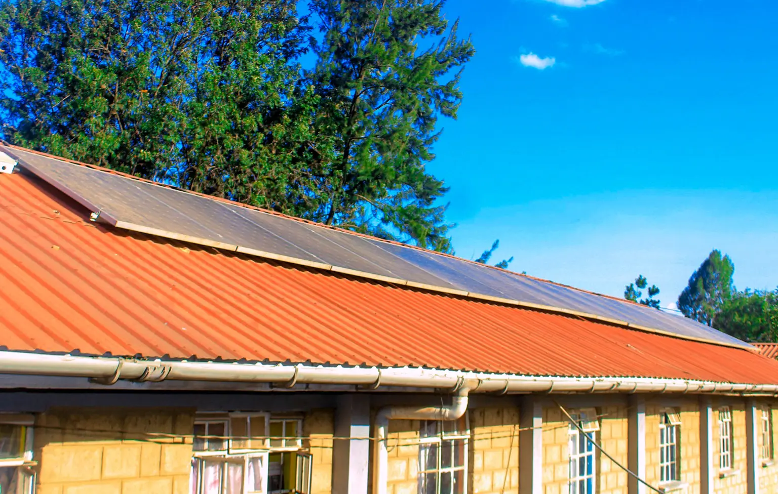 Solar panels installed on a red, corrugated metal roof of a single-story building under a clear blue sky. Trees are visible in the background.