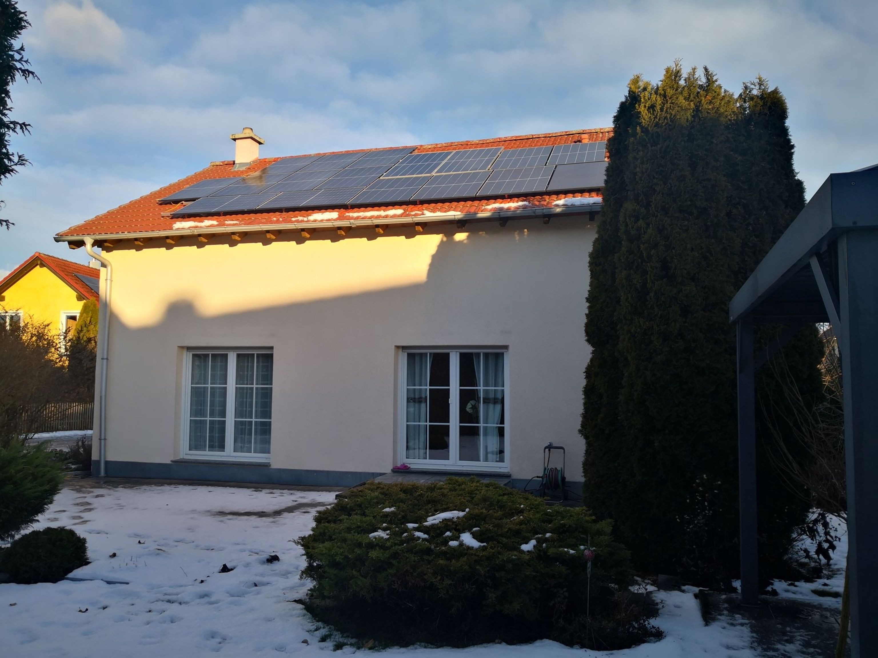 A house with solar panels on the roof, surrounded by snow, bushes, and a tall evergreen tree under a partly cloudy sky.