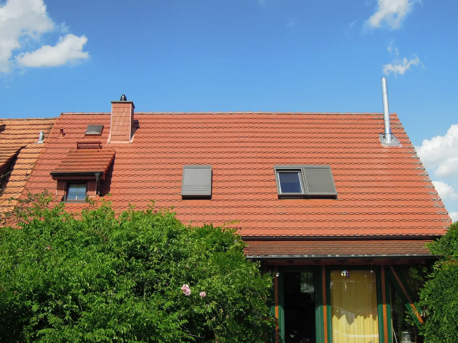 Viewed from the garden, the house has a red roof with a skylight, green-framed windows and a balcony door, with a large bush in front.