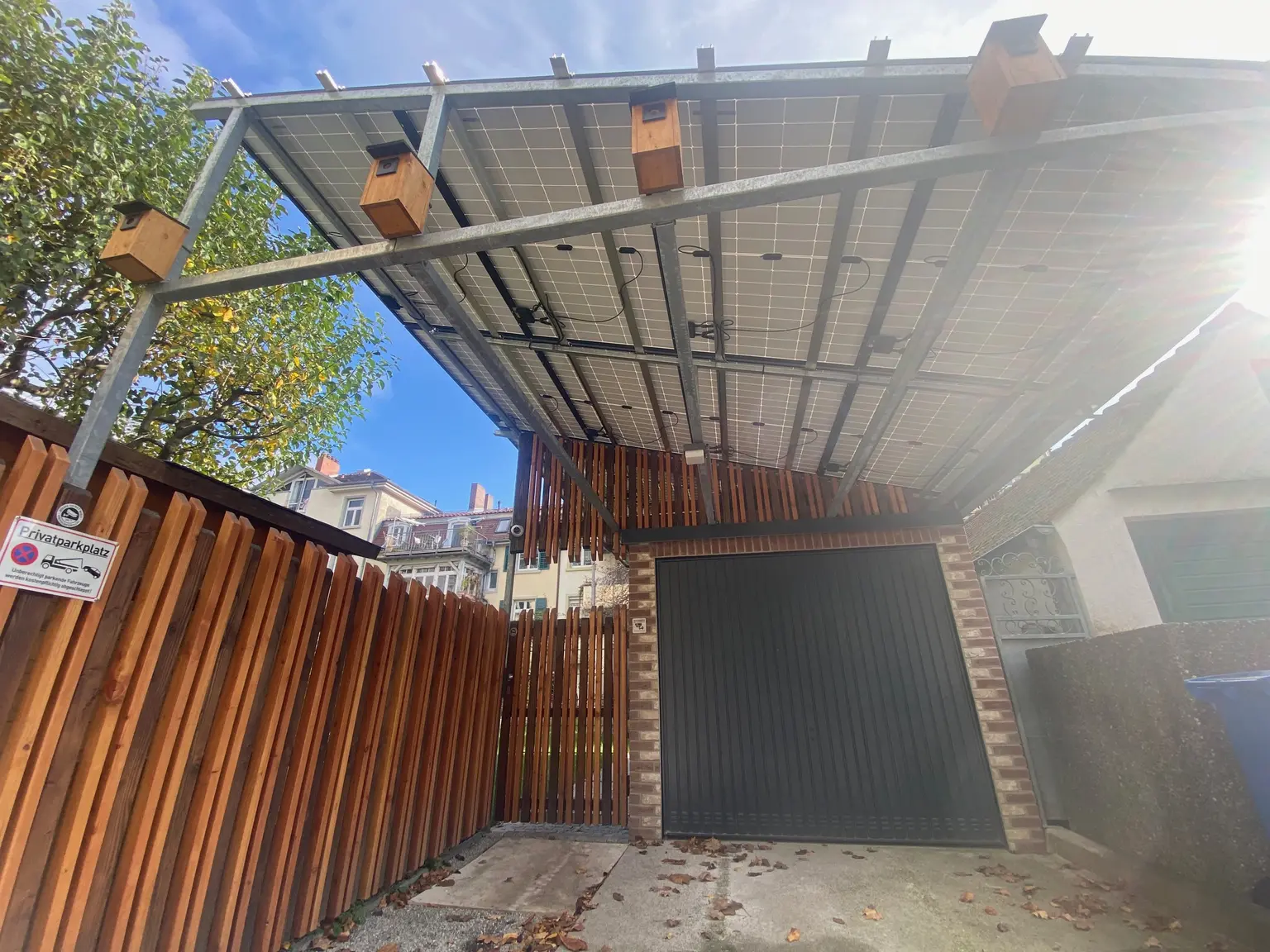 Driveway to a garage, surrounded by a fence and covered with a photovoltaic system.