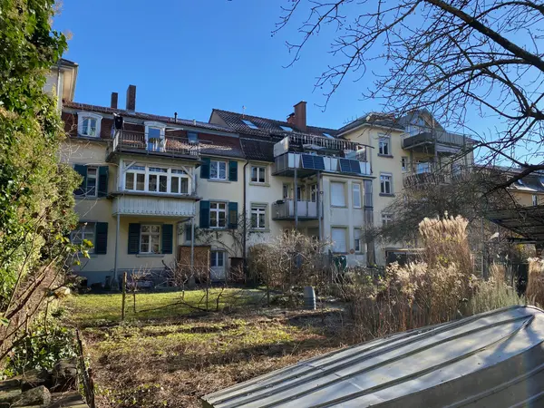 Rear view of a multi-story residential building with balconies, surrounded by a garden and trees under a clear blue sky.