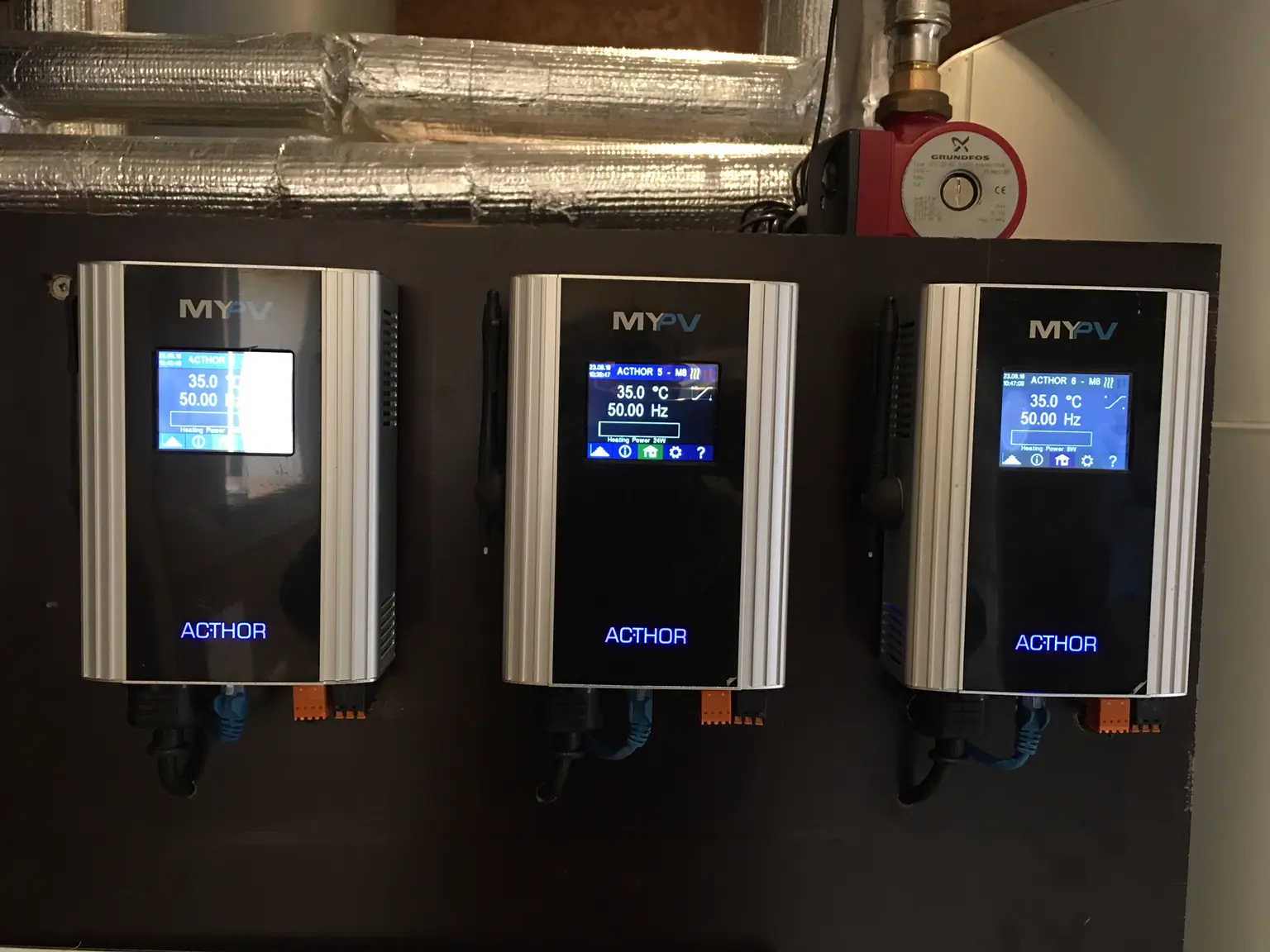 Three my-PV AC•THOR devices mounted on a wall, displaying digital screens with temperature and power settings. Pipes are visible above.