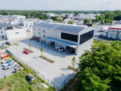 Aerial view of a modern industrial building with solar panels, surrounded by paved parking and greenery in a suburban setting.