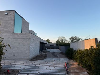 Modern building exterior with large glass windows, surrounded by trees. The pathway is lined with white rectangular tiles.