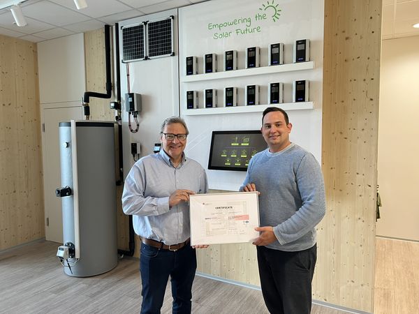 Two men holding a certificate stand in a room with solar equipment and a sign that reads "Empowering the Solar Future."