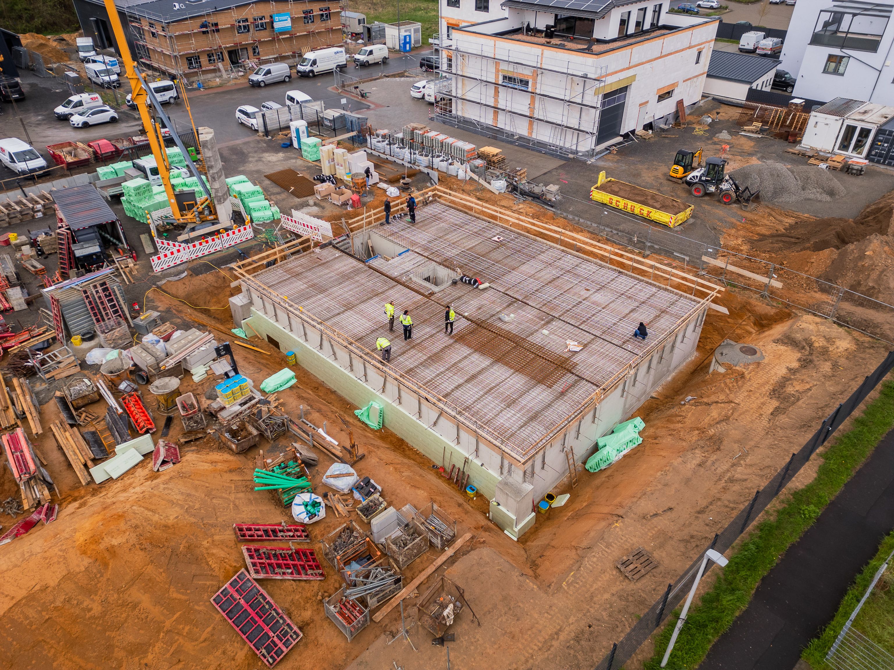 Aerial view of a construction site with workers on a concrete foundation, surrounded by materials, machinery, and partially built structures.