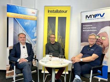 Three men seated around a small table with glasses, surrounded by promotional banners.
